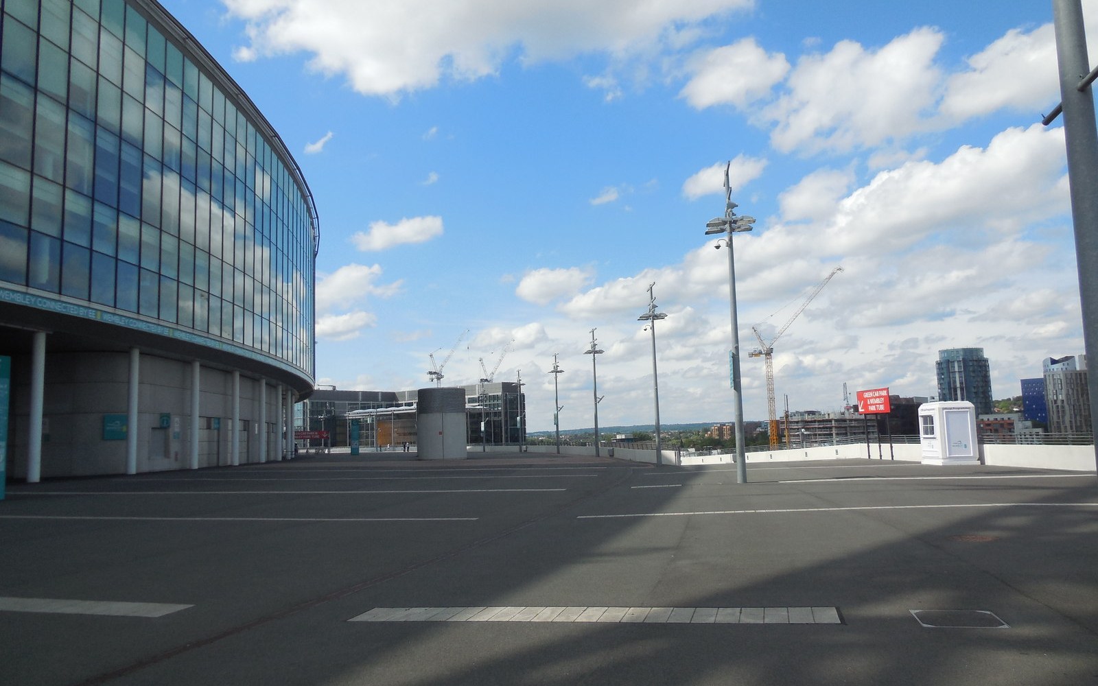 Empty parking area beside a curved glass stadium facade, with cranes on the skyline